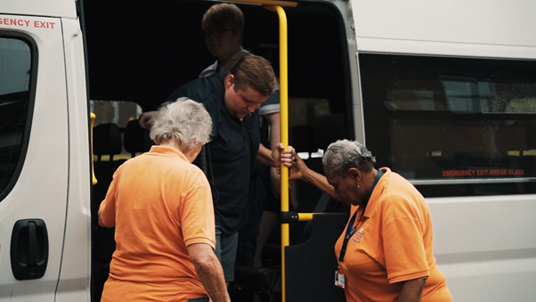 http://A%20young%20man%20steps%20down%20from%20a%20minibus%20while%20two%20Bradbury%20Fields%20staff%20members%20in%20orange%20shirts%20stand%20nearby%20to%20offer%20support.