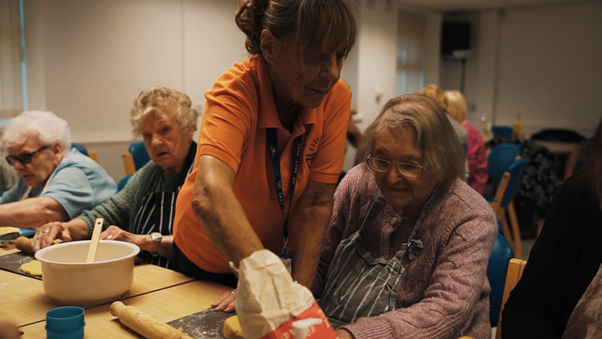 http://A%20Bradbury%20Fields%20staff%20member%20supports%20an%20older%20woman%20with%20rolling%20dough%20during%20a%20group%20baking%20activity,%20with%20other%20participants%20seated%20around%20the%20table.