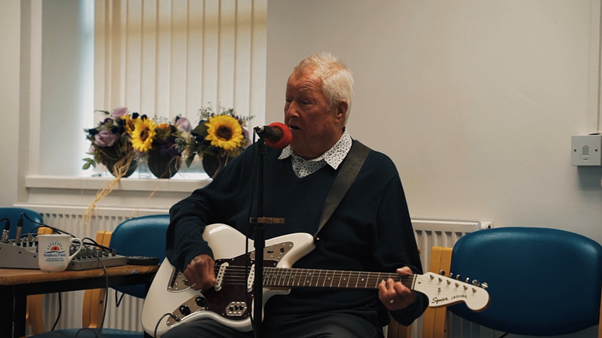 http://An%20older%20man%20sits%20playing%20an%20electric%20guitar%20and%20singing%20into%20a%20microphone%20during%20a%20social%20activity%20at%20Bradbury%20Fields.