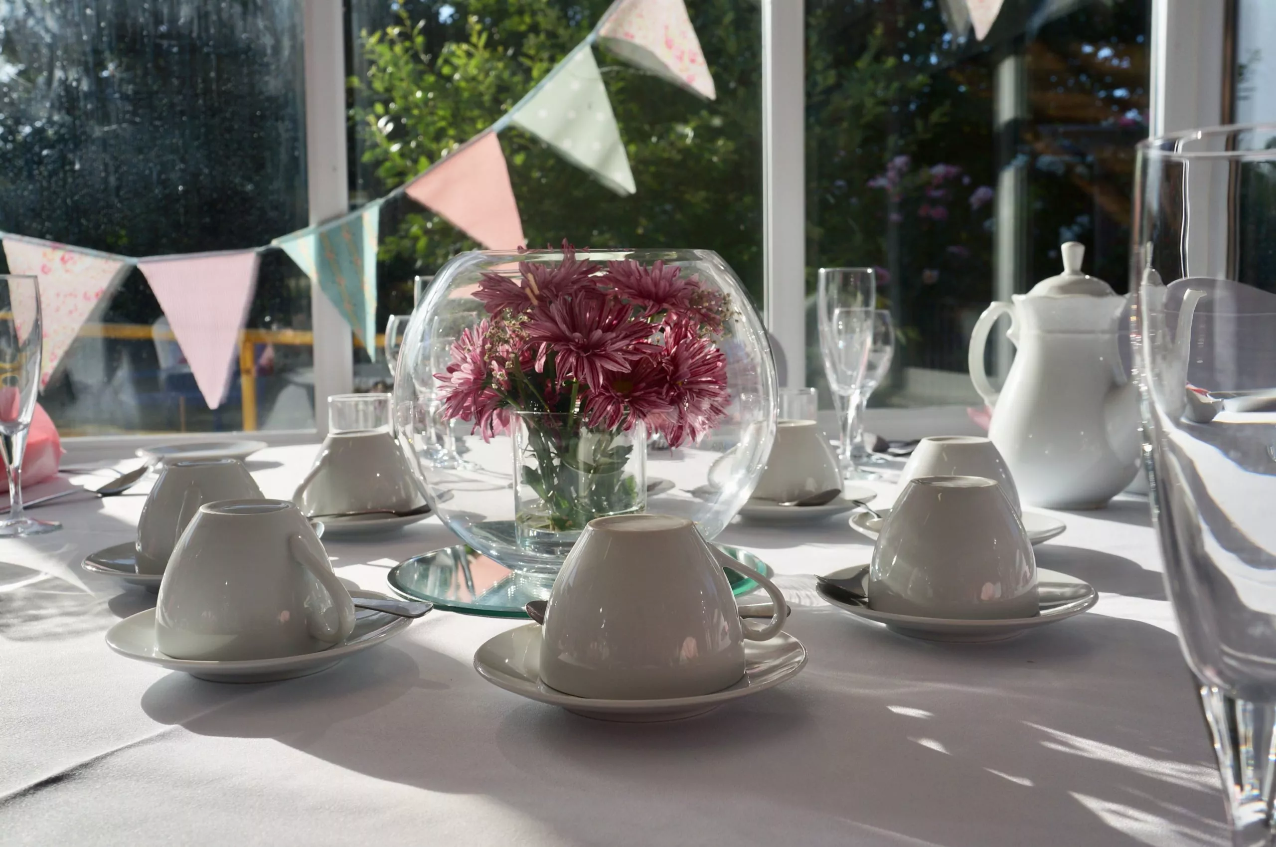 http://A%20close-up%20of%20an%20afternoon%20tea%20table%20setting.%20The%20sun%20is%20shining%20through%20the%20glass%20centrepiece%20filled%20with%20pink%20flowers%20and%20there%20is%20bunting%20and%20a%20view%20of%20the%20sensory%20garden%20in%20the%20background.